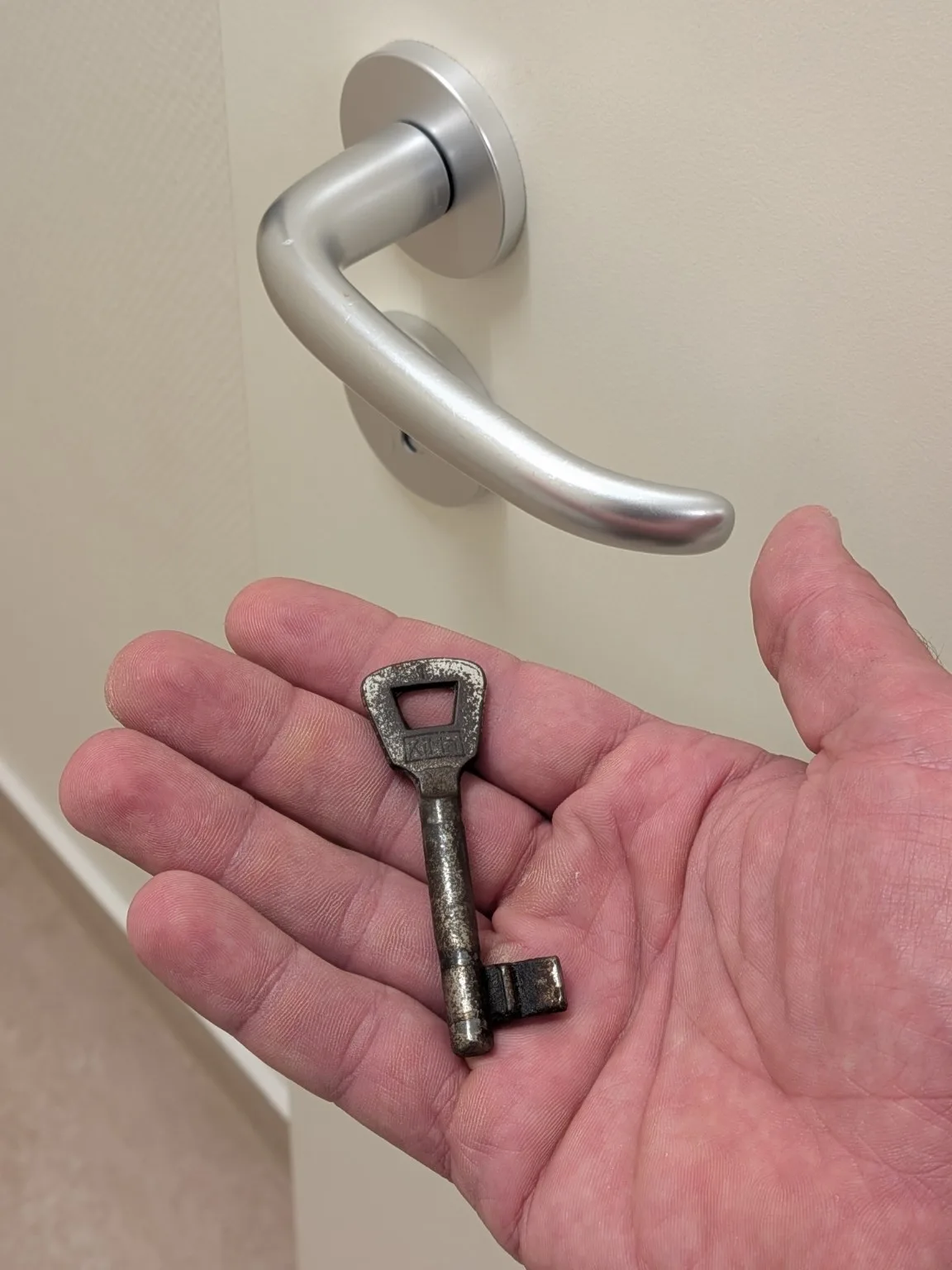 Gebrauchter Bartschlüssel in einer Hand vor einer Tür mit silbernem Drücker. Gebrauchsspuren sagen allein wenig über Funktion und Einbruchschutz des Schlosses aus.